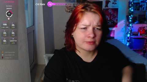 Media: A video of a pale-skinned woman with dyed red hair, wearing a black t-shirt, sitting indoors, surrounded by gaming-themed decor and digital overlays.