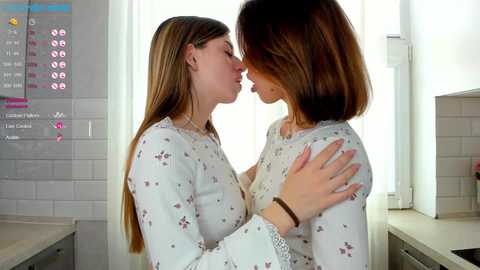 Media: Video of two young women in white floral-patterned nightgowns, embracing passionately in a bright, modern kitchen with white tiles and a digital menu screen visible on the wall.