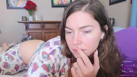 Media: Video of a young woman with light skin and brown hair, wearing colorful pajamas, lying on a bed, touching her lips, in a bedroom with framed art and a vase of flowers.