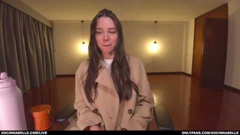 Media: Video of a young woman with long brown hair, wearing a beige trench coat, sitting at a wooden table in a dimly lit room with beige walls and a brown floor.