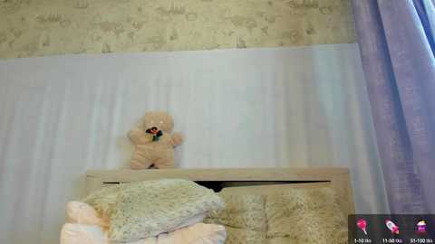 Media: Video of a teddy bear on a bed, wearing a green bow tie, in a room with light wallpaper and a purple curtain.