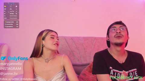 Media: Video of a young woman with long brown hair, wearing a white lace dress, and a man with glasses, black shirt, and mustache, sitting on a couch under pink lighting.