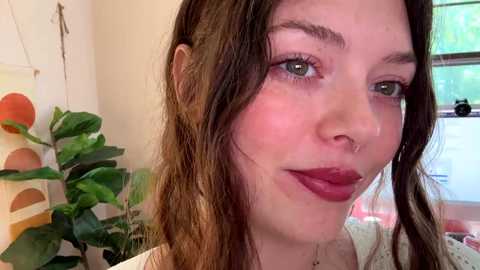 Media: Video of a young woman with fair skin, green eyes, and brown hair, wearing a nose ring and red lipstick, smiling. Background includes a potted plant and a window with greenery outside.