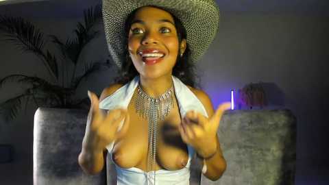 Media: Video of a joyful Black woman with curly hair, wearing a cowboy hat, silver necklace, and a white halter top, exposing her breasts, in a dimly lit room with a gray couch and potted plants.