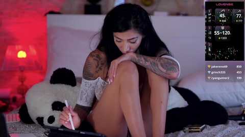 Media: Video of a tattooed, dark-haired woman with a slender physique, wearing a white lace top, sitting on a bed with a panda plush, while holding a pen, in a dimly lit room.