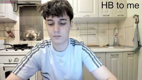 Media: Video of a young boy with messy hair, wearing a white Adidas t-shirt, sitting in a modern kitchen with white cabinets, stove, and utensils. Text reads \"HB to me.\