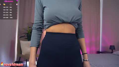 Media: Video of a woman in a grey crop top, high-waisted black leggings, standing in a bedroom with a bed, potted plant, and pink lighting.