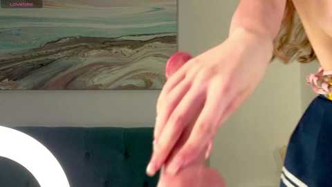 Media: Video of a woman's hand holding a large, erect penis, in a modern, minimalistic room with a marble-patterned painting and a teal couch.