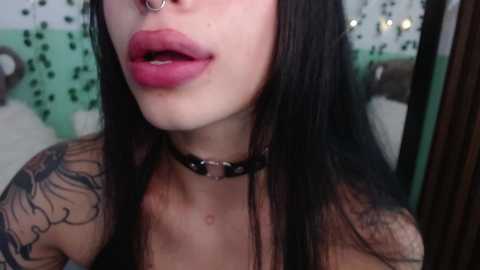 Media: Video of a pale-skinned woman with long black hair, full lips, a nose ring, and a black choker, featuring a tattooed arm in a teal and green bedroom.