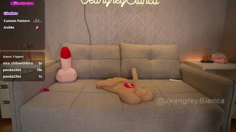 Media: Video of a cozy bedroom with a plush, beige sofa featuring a large, realistic, flesh-toned dildo and a pink plush toy. The background shows a patterned wall with a glowing \"Welcome Home\" sign.