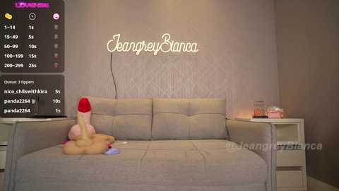 Media: A video of a beige couch in a dimly lit room with a large, pink dildo and a stuffed toy on it. The wall behind features a \"Jasmine Bana\" sign and two white nightstands with lamps.