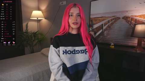 Media: Video of a young woman with long, bright pink hair, wearing a gray and black \"Hoodies\" sweatshirt, standing in a dimly lit bedroom with a TV playing a beach scene.