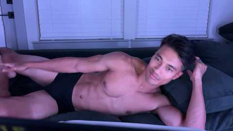 Media: Video of a shirtless Asian man with short black hair lying on a dark bed, wearing black briefs, under white blinds.