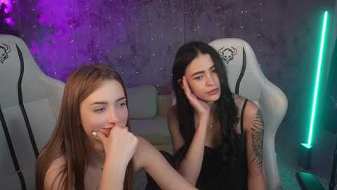 Media: Video of two young women with fair skin, one with long brown hair and the other with long black hair, sitting in gaming chairs with a purple and green neon backdrop.