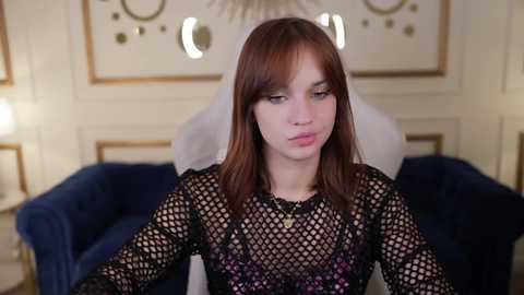 Media: Video of a young woman with long brown hair, wearing a black fishnet top, sitting on a plush blue couch in a luxurious, dimly lit room.