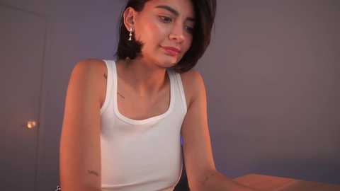 Media: Video of a young woman with short, dark hair, wearing a white tank top, standing indoors against a muted purple backdrop, with soft, warm lighting.