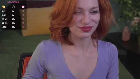 Media: Video of a fair-skinned woman with shoulder-length red hair, wearing a purple V-neck top, sitting in an orange chair. Background shows a green wall with a plant and a yellow couch.