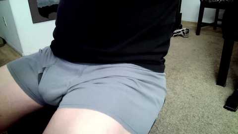 Media: A video of a person sitting on a beige carpet in a room, wearing grey shorts that are slightly tight and a black shirt. The background includes a black chair and white shoes on the floor.