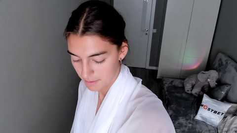 Media: A video of a young woman with dark hair in a white robe, standing in a modern bedroom with a gray carpet, a gray bed, and a plush gray teddy bear.