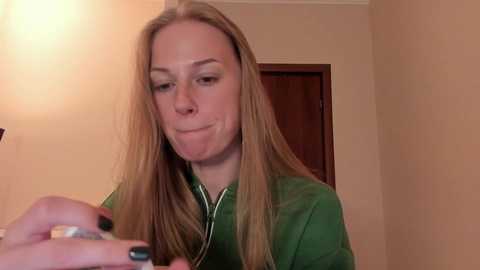 Media: A video of a Caucasian woman with long, straight, blonde hair, wearing a green zip-up jacket, sitting indoors, focusing on a small object in her hands.