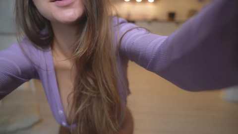 Media: A video of a young woman with long, straight brown hair, wearing a purple, ribbed cardigan, and smiling slightly. The background is a blurred, warmly lit indoor setting.