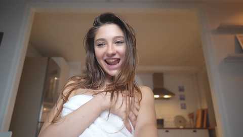 Media: Video of a smiling, topless woman with wet, long brown hair in a modern kitchen, wearing a white towel, holding it against her chest.