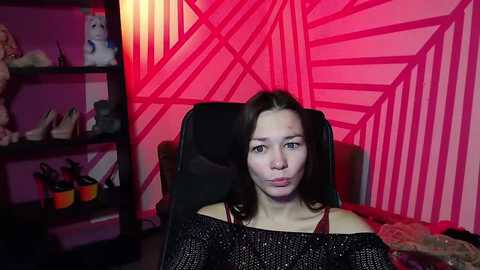 Media: Video of a young woman with fair skin and dark hair, wearing a black off-shoulder sweater, sitting in a black chair against a pink geometric-patterned wall. The room is dimly lit with shelves filled with toys and shoes in the background.