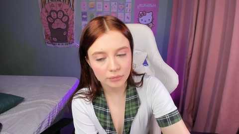 Media: A video of a young Caucasian woman with fair skin and long auburn hair, wearing a plaid shirt, sitting in a white gaming chair in a cozy, pink-draped bedroom.