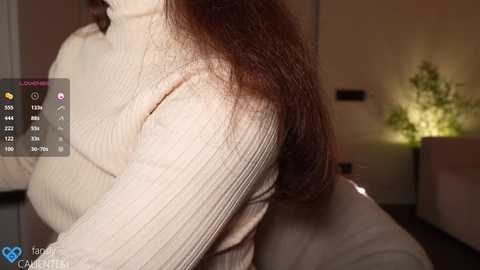 Media: A close-up video of a woman with long, straight brown hair, wearing a cream ribbed turtleneck sweater. Background features a cozy, dimly-lit room with a potted plant and soft lighting.