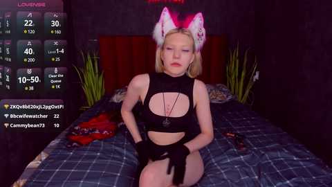 Media: Video of a blonde woman in a black, cut-out bodysuit, with a cat ear headband, sitting on a bed in a dimly-lit room, with a digital screen displaying streaming stats.