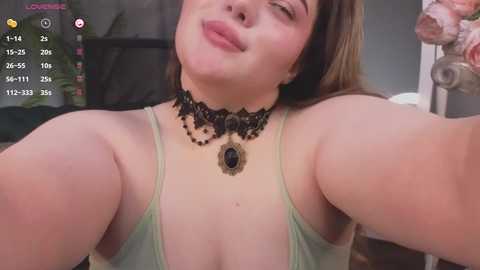 Media: A video of a light-skinned woman with long brown hair, wearing a low-cut, light green spaghetti strap top, black choker with a large pendant, and a playful, slightly puckered mouth.