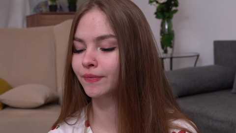 Media: Video of a young woman with fair skin and long brown hair, wearing a white shirt with a floral pattern, sitting on a beige couch in a living room with a gray armchair and a green plant.