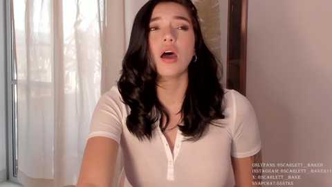 Media: Video of a young Asian woman with long black hair, wearing a white t-shirt, looking surprised or shocked with parted lips, indoors with white curtains.