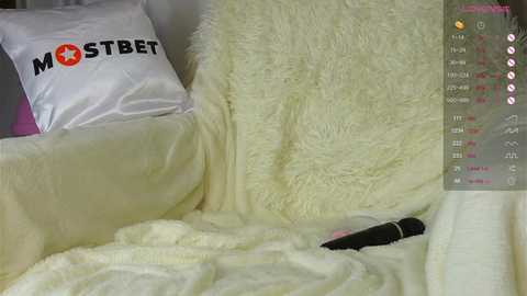 Media: Video of a plush, white, faux fur blanket covering a bed with a black vibrator on it. The background features a white pillow with \"MOSTBET\" text. The scene has a casual, intimate atmosphere.