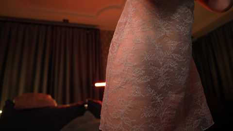 Media: A video of a woman's bare upper torso in a dimly lit bedroom, wearing a sheer, floral-patterned lace nightgown, with heavy curtains in the background.