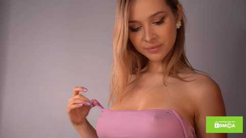 Media: Video of a light-skinned, blonde-haired woman with medium-sized breasts, wearing a strapless pink dress, adjusting a strap, in a simple indoor setting.