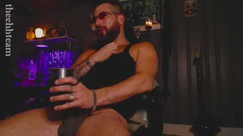 Media: Video of a muscular, bearded man in a black tank top, holding a glass, seated indoors with dim lighting, tattoos, and a skull on the wall.