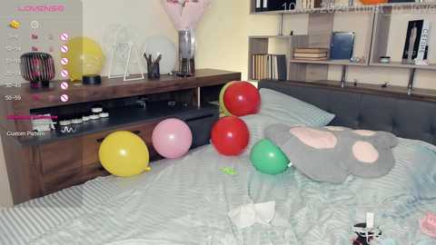 Media: Video of a modern bedroom with a bed covered in light blue striped bedding. The room features a wooden bedside table with a vase, pens, and books. Five colorful balloons (yellow, pink, green, red, and gray) are scattered on the bed.