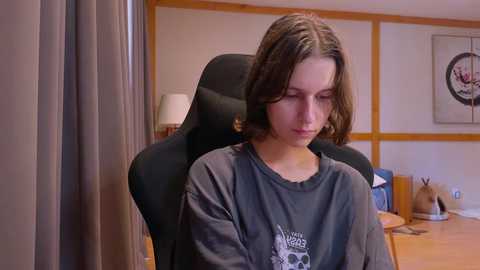 Media: Video of a young woman with shoulder-length brown hair, wearing a gray \"The Great Gatsby\" T-shirt, sitting in a black office chair in a modern office with wooden paneling and a framed circular logo.