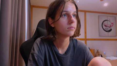 Media: Video of a young woman with shoulder-length brown hair, wearing a dark gray t-shirt, sitting in a black chair in a modern, wooden-paneled room with a framed abstract painting on the wall.