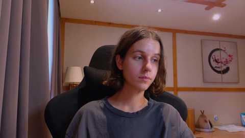 Media: Video of a young girl with light skin and shoulder-length brown hair, sitting in a black office chair, wearing a gray sweater. Background features wooden paneling, a lamp, and a framed painting.