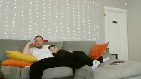 Media: Video of a man and woman lying on a gray couch, watching TV; the woman is asleep with her head on the man's shoulder. Background features a string of fairy lights and an orange throw pillow.