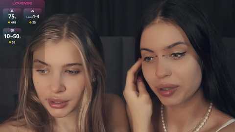 Media: Video of two young women, one blonde with fair skin, the other brunette with olive skin, both wearing makeup, sitting close, in a dimly lit room with a dark background.