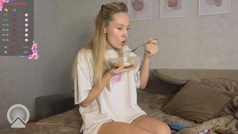 Media: A video of a young blonde woman in a white T-shirt and shorts eating cereal from a bowl on a bed with brown sheets.