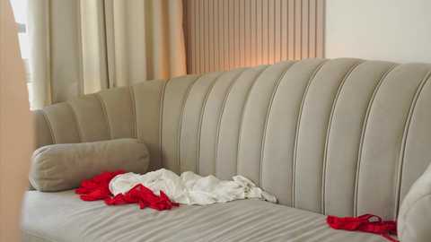 Media: Video of a modern beige sectional sofa with vertical tufted cushions. A red and white striped dress is casually draped over the backrest. Cream-colored curtains and a wooden panel backdrop provide a soft, minimalist setting.