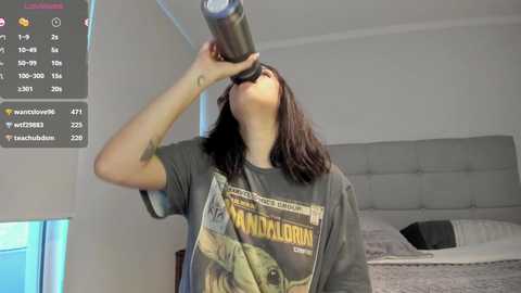 Media: Video of a woman in a dark T-shirt with a Star Wars design, drinking from a silver can, standing in a modern bedroom with a bed and a digital clock showing 4:51 AM.