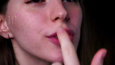 Media: Close-up video of a young woman with fair skin, brown hair, and freckles. She has a finger to her lips, indicating shush, with a subtle, pinkish glow on her skin.