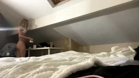 Media: Video of a blonde woman in a white bra and panties adjusting a fluffy blanket on a bed in a dimly lit attic bedroom with a sloped ceiling.