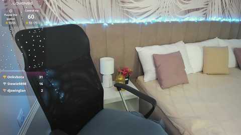 Media: Video of a modern bedroom with a white headboard, beige walls, and a light blue string of fairy lights. A black office chair is prominently placed in the foreground, and a small nightstand holds a pink cushion.