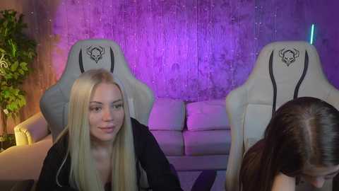 Media: Video of two women, one blonde and one brunette, sitting on white gaming chairs with a skull logo, in a dimly lit room with purple and blue lighting.